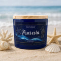 Vela artesanal Maresia Solenza em pote de vidro azul com tampa de madeira, apoiada na areia com conchas e mar ao fundo.